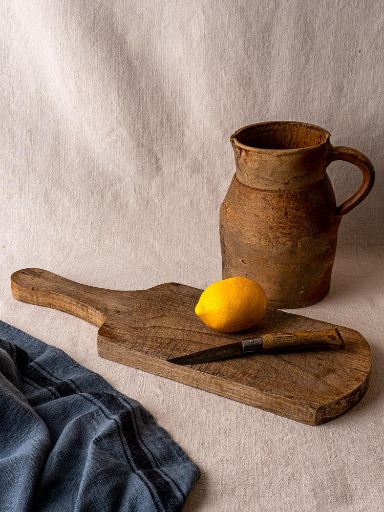 Antique French oak and chestnut cutting board with lemon, knife, and vintage ceramic jug on neutral cloth.