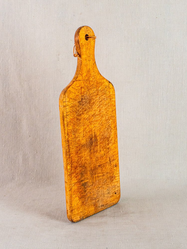 Antique French cutting board with paddle handle and rounded edges, worn wood against neutral fabric backdrop.