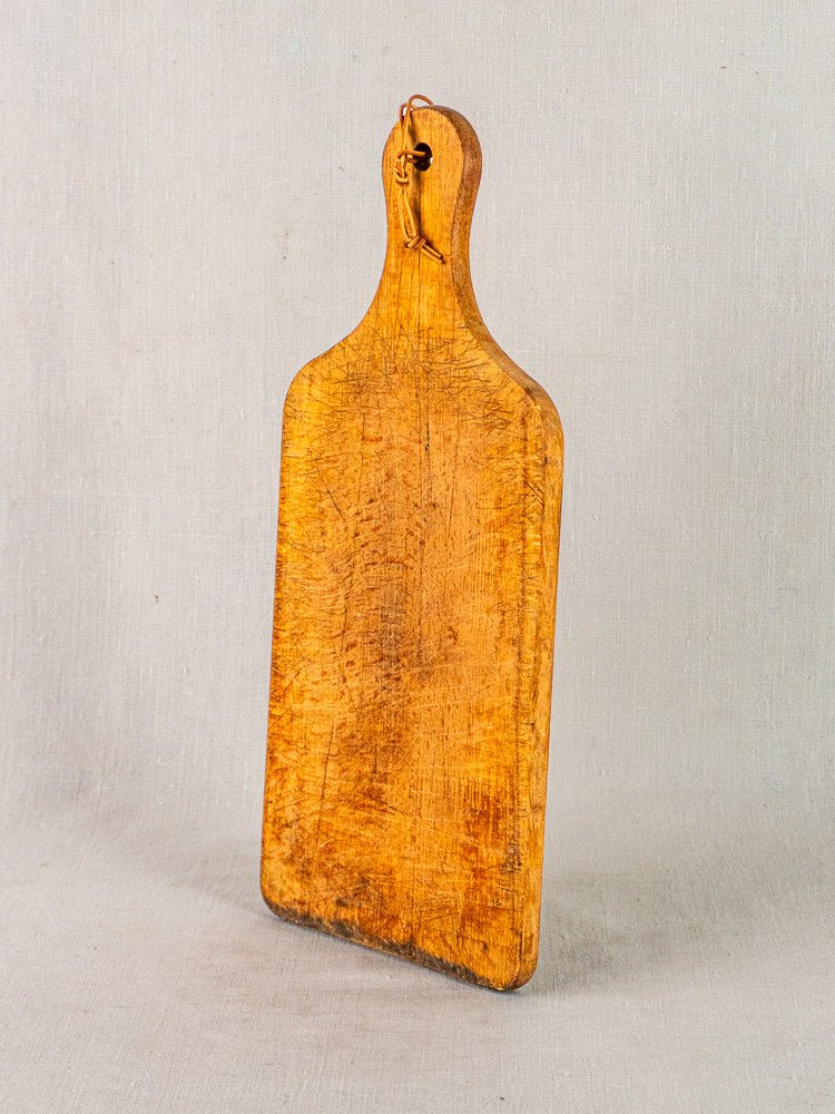 Antique French wooden cutting board with paddle handle and leather loop, upright on neutral background.