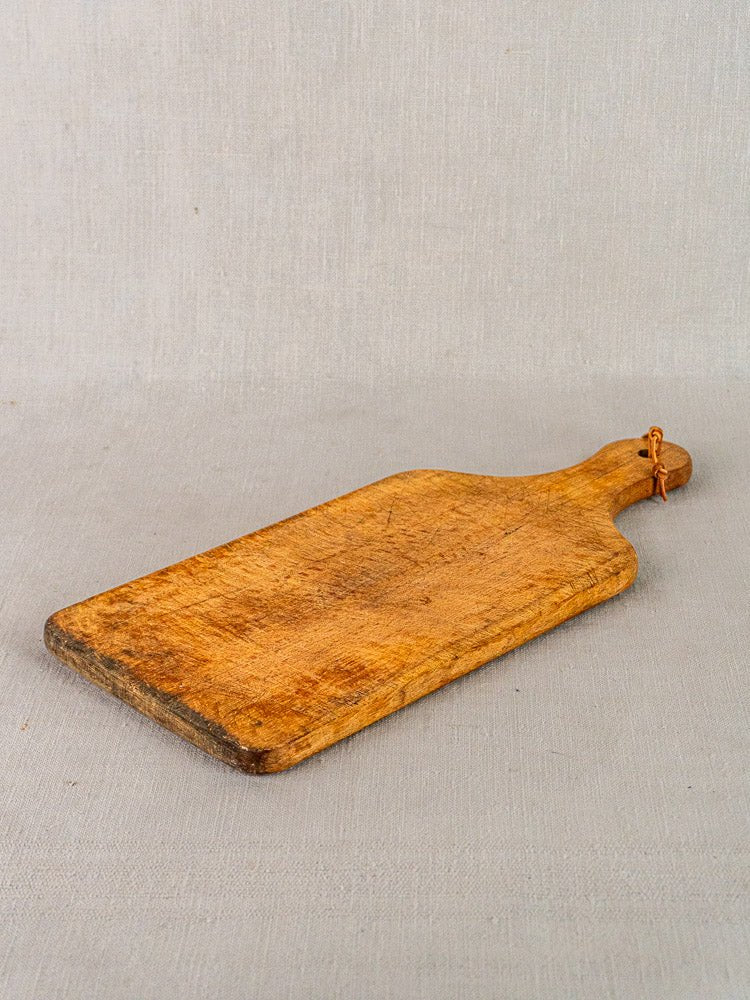 Antique French rectangular wooden cutting board with paddle handle and hanging loop on light gray surface.