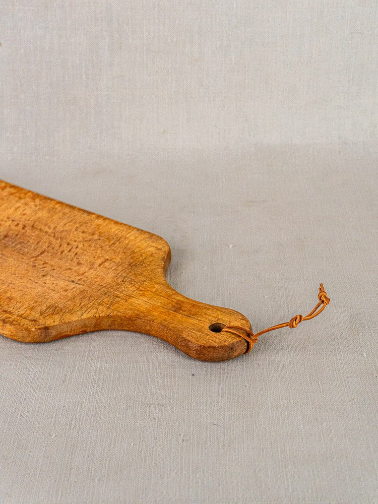 Antique French cutting board with paddle handle and twine loop on light gray fabric surface.