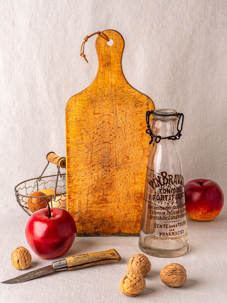 Antique French cutting board with paddle handle, vintage glass bottle, apples, walnuts, eggs in wire basket.