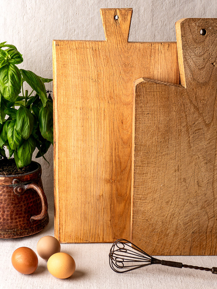 Antique French cutting board with carved paddle handle, styled with basil plant, whisk, and eggs on a light surface.