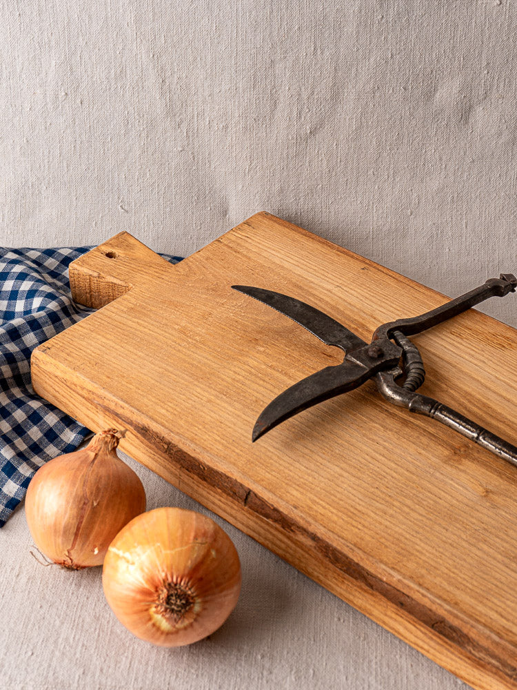 Antique French cutting board with carved paddle handle, early 20th century, beside onions and checkered cloth.