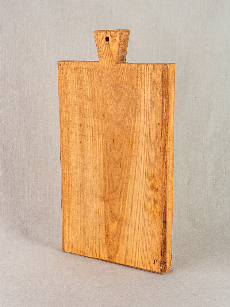 Early 20th-century antique French cutting board with carved paddle handle, upright against neutral fabric background.