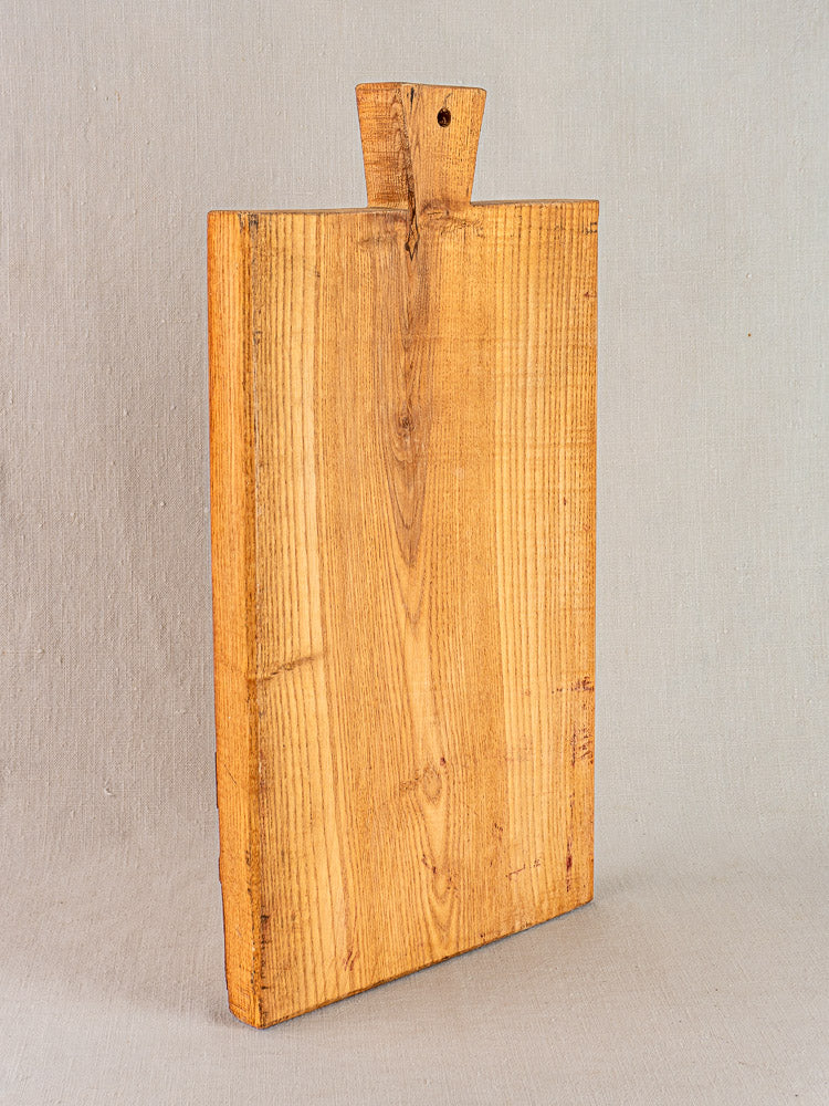 Antique French cutting board with carved paddle handle and hanging hole, showcasing rich wood grain on neutral fabric.