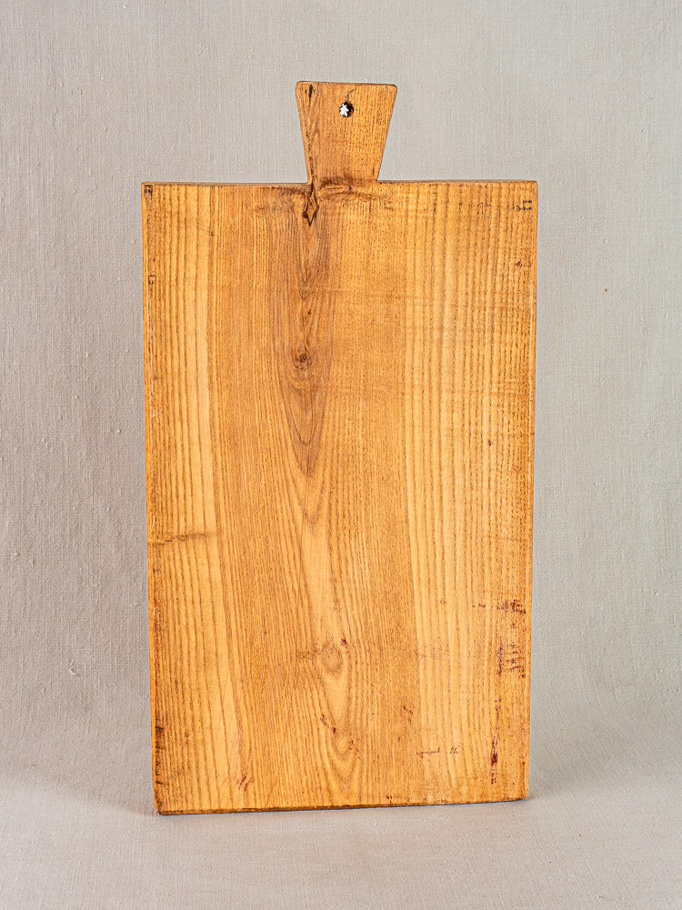 Antique French cutting board with carved paddle handle, early 20th century, upright on light gray fabric background.