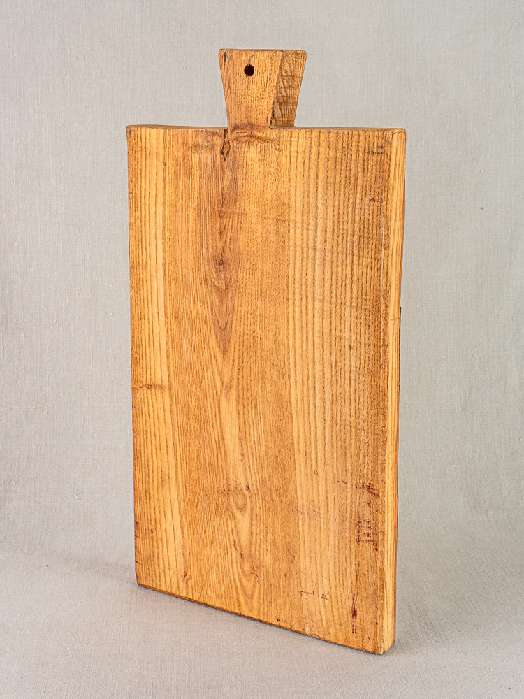 Antique French cutting board with carved paddle handle and hanging hole, showcasing wood grain on a light surface.