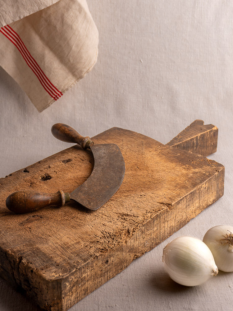 Antique French cutting board with curved herb chopper, styled with white onions and beige cloth for timeless farmhouse decor.