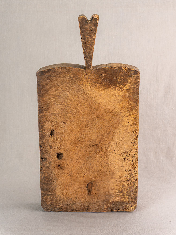 Antique French cutting board from the 1880s with heart-shaped handle and weathered patina, upright against a plain background.