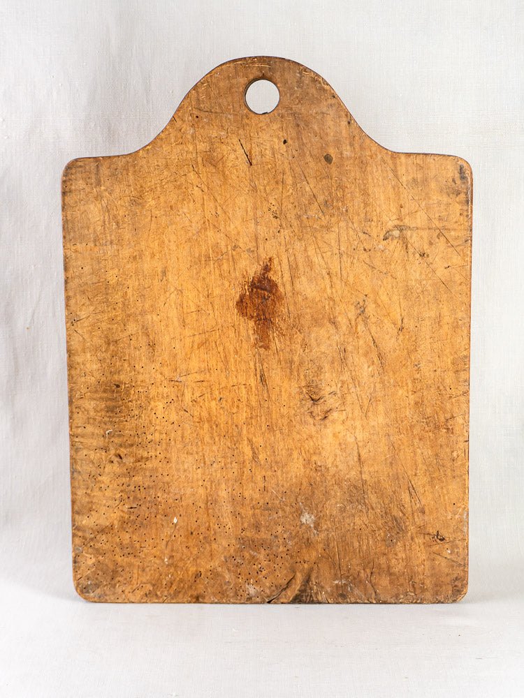 Antique circa 1900 French beech chopping board with rounded top and central hole, upright on light background.