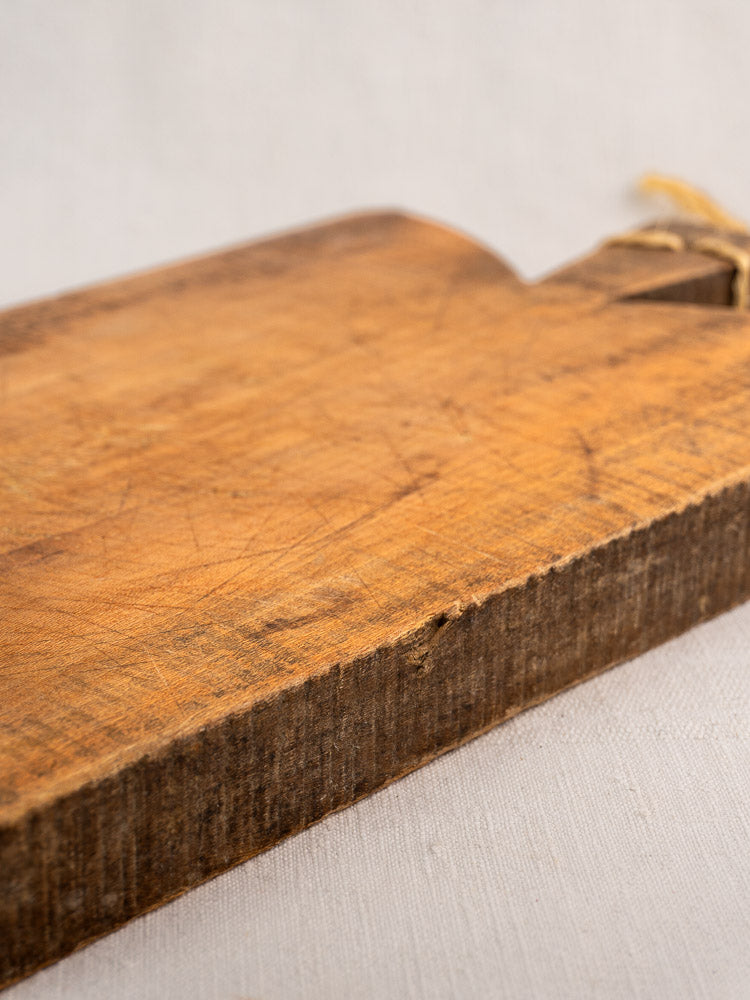 Antique French cutting board with textured, scratched surface and rope-tied handle on light fabric.