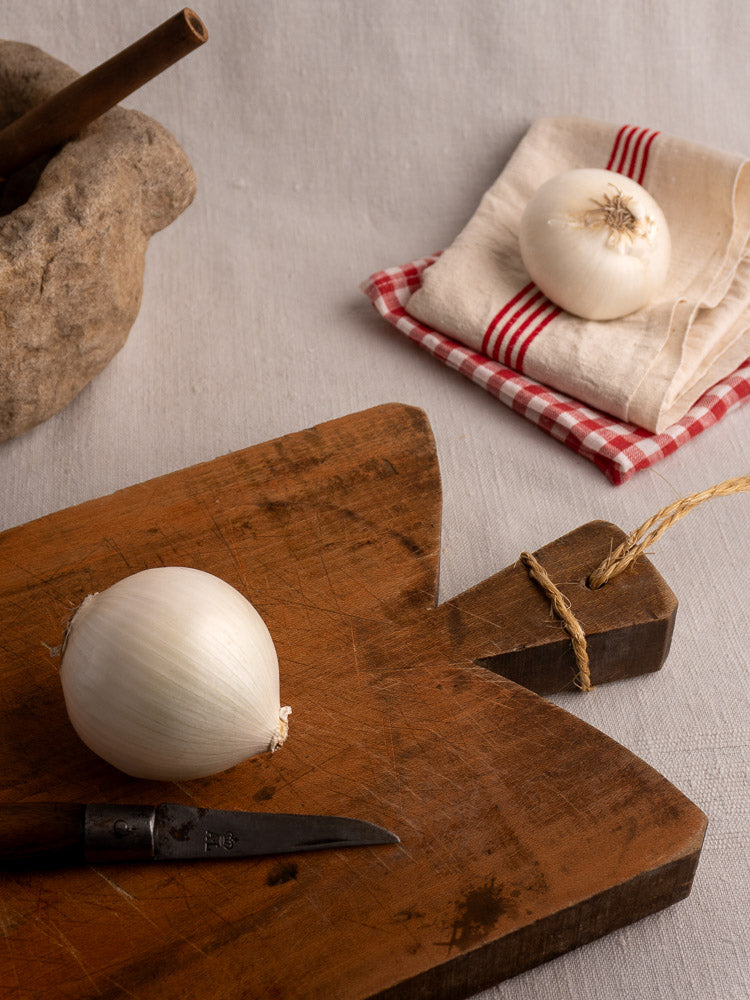 Antique French single plank cutting board with white onion, small knife, and stone mortar and pestle on white linen.