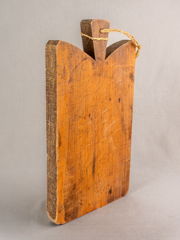 Antique French cutting board with handle and twine, rough edges, and knife marks set against a neutral background.