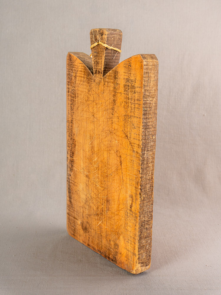 Antique French single plank cutting board with rustic knife marks, upright on a neutral gray surface, handle tied with string.