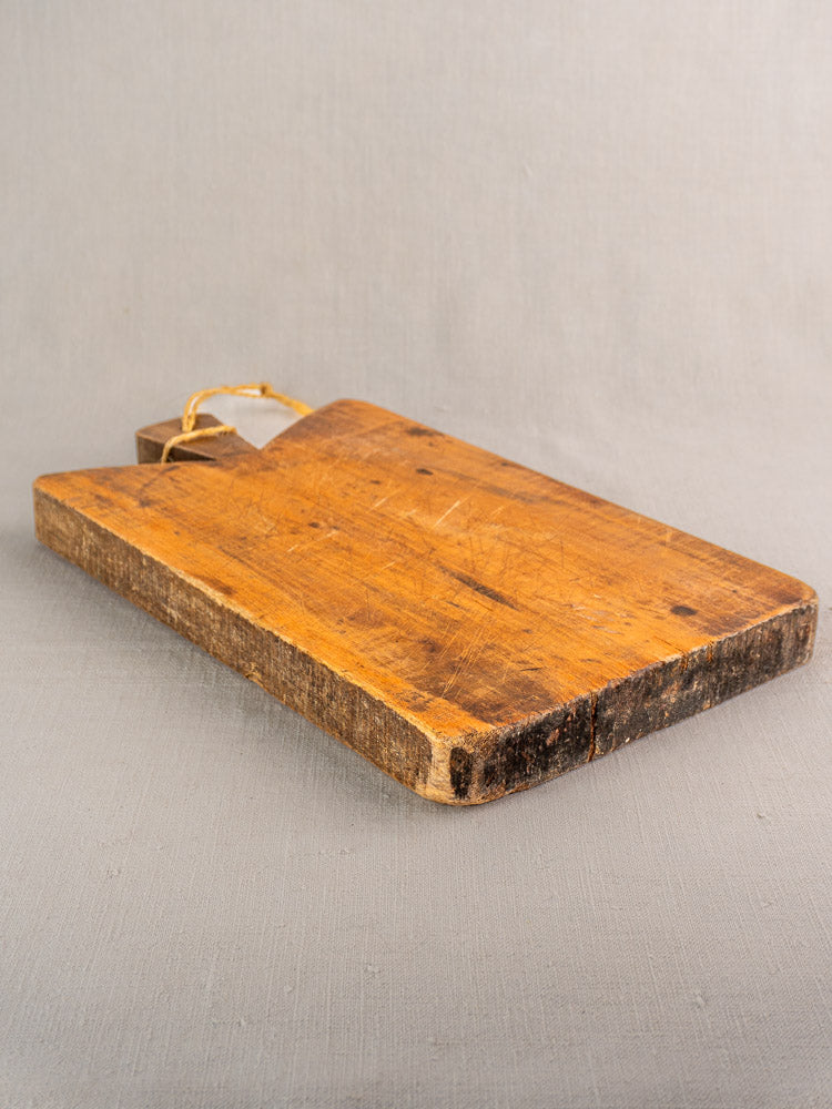 Antique French cutting board, late 19th century, rectangular with handle and string loop, heavily worn, on a gray surface.