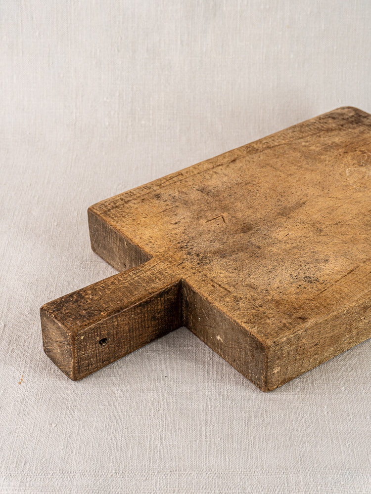 Antique French cutting board with tongue handle, visible grain and wear, on a light beige fabric surface.