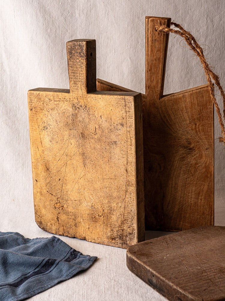 Antique French cutting board with tongue handle, vintage grain and wear, upright on light fabric, folded blue cloth beside it.
