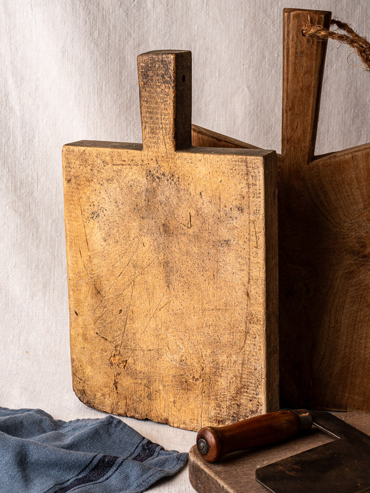Antique French cutting board with tongue handle, showing age marks, upright on neutral fabric background.