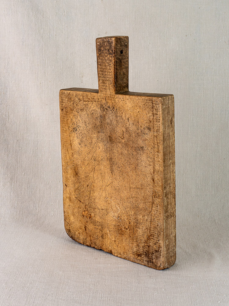 Antique French cutting board with tongue handle, showing age marks, upright on neutral fabric background.
