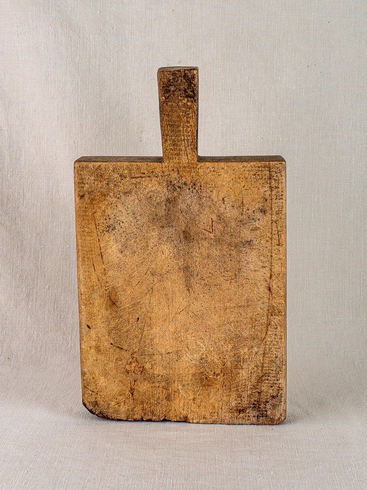 Antique French cutting board with tongue handle, circa 1900, upright showing scratches and discoloration against a light textured background.