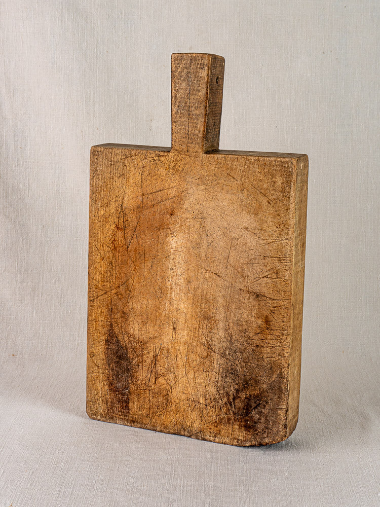 Antique French cutting board from circa 1900 with tongue handle and knife marks, shown upright on light fabric.