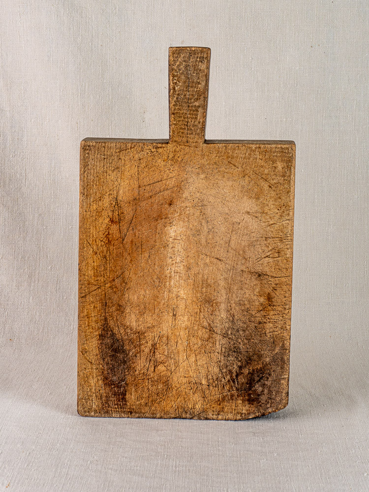 Antique French cutting board with tongue handle, vertical against light textured fabric, worn with age-darkened marks.