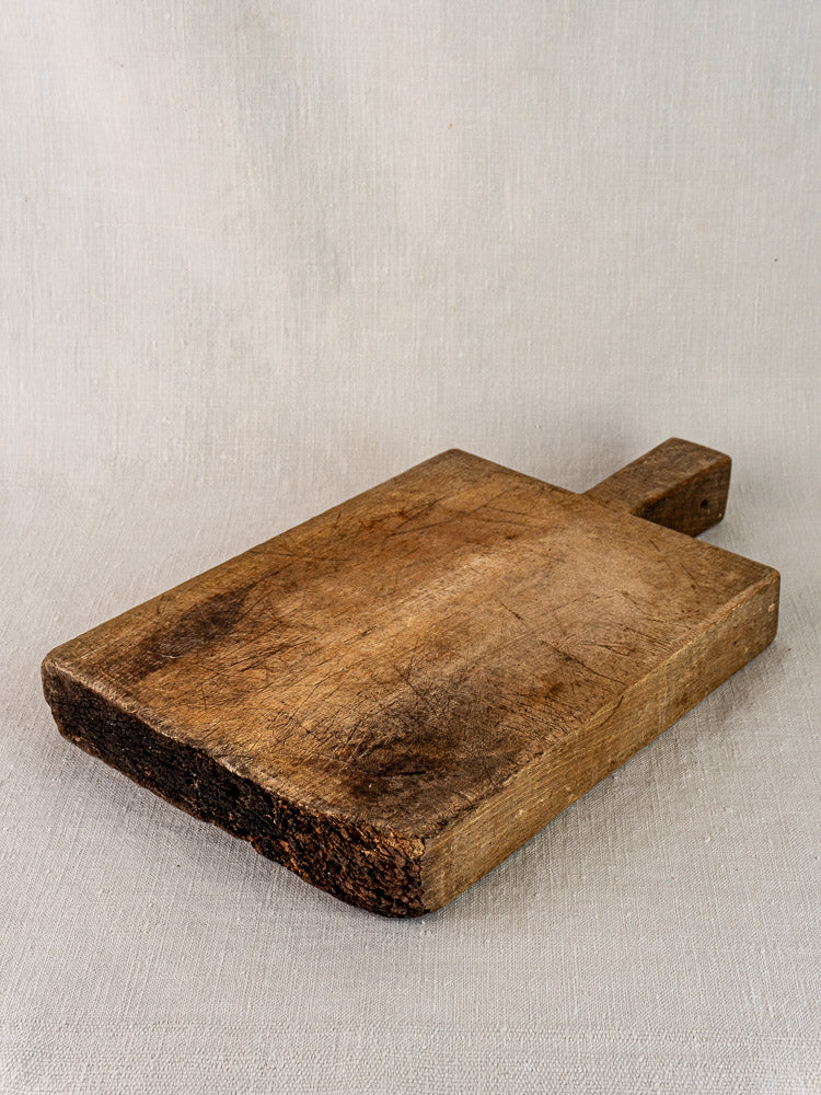 Antique French cutting board from solid wood with tongue handle, worn edges and surface scratches on a light textured background.