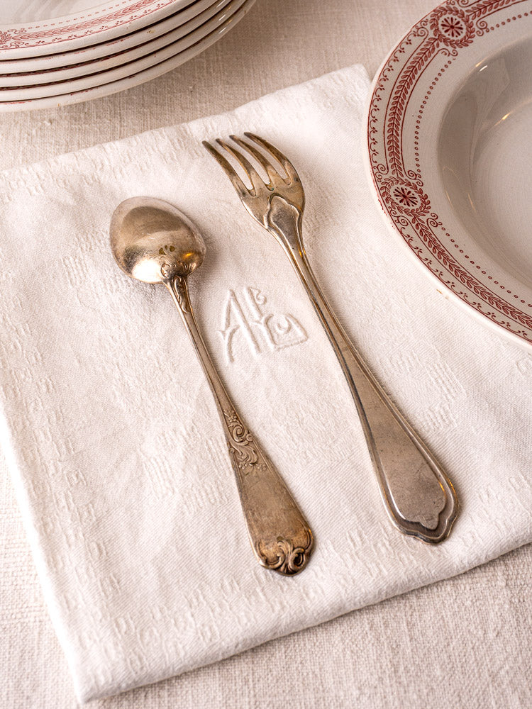 Antique French monogrammed damask linen napkins with vintage silverware beside patterned bowl and red-trimmed plates.
