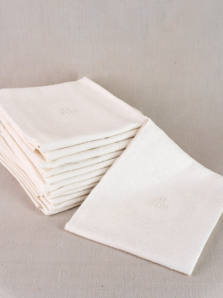 Set of 12 antique French damask linen napkins, each monogrammed and neatly folded on a neutral fabric background.