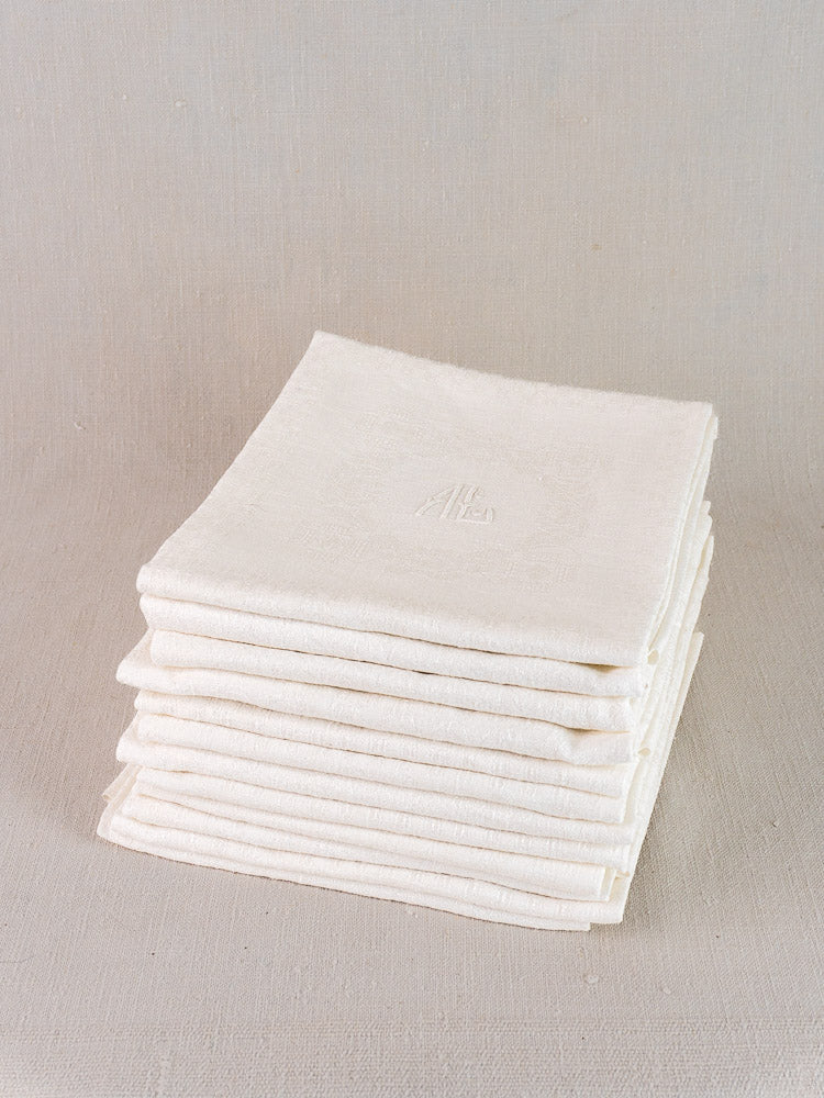 Set of 12 antique French damask linen napkins, each monogrammed, stacked on a light beige surface.