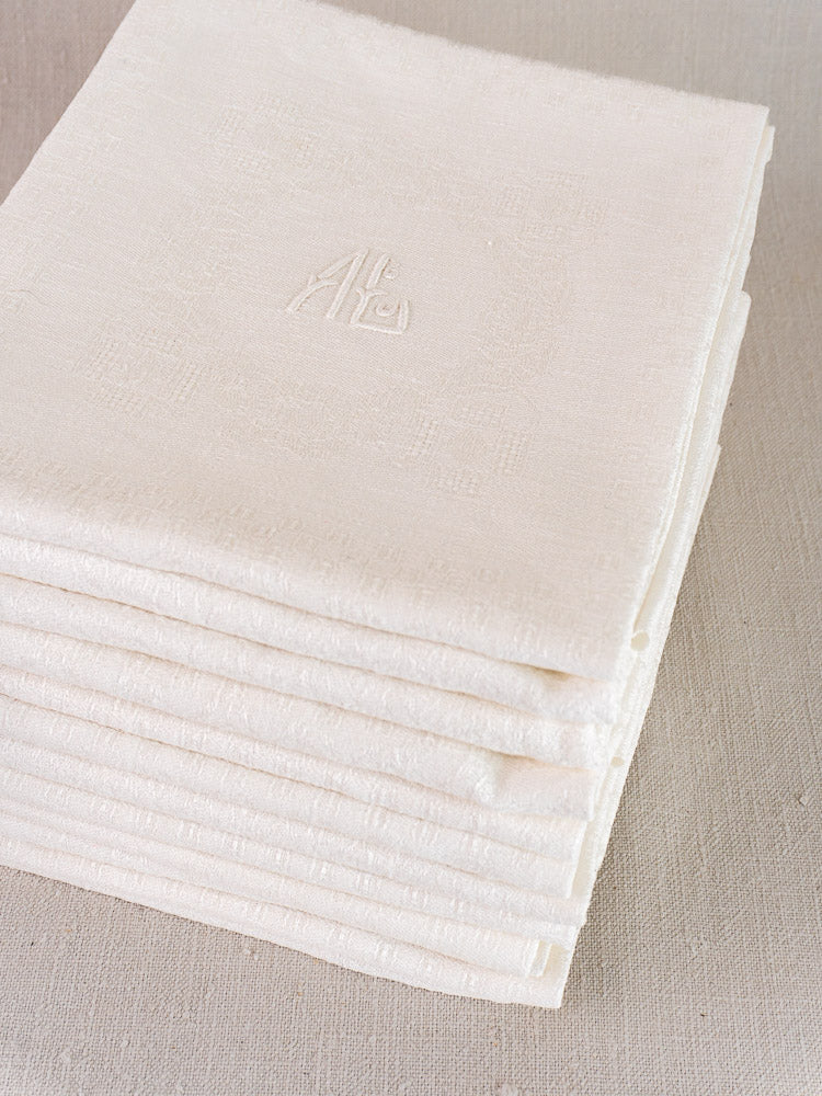 Set of 12 antique French damask linen napkins, each with delicate monogram embroidery, on a light fabric background.