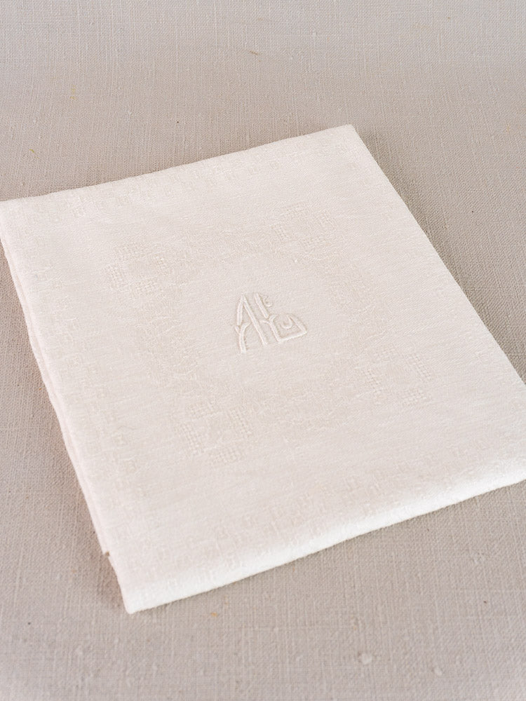 Antique French damask linen napkin with monogram, neatly folded on a light beige fabric surface.