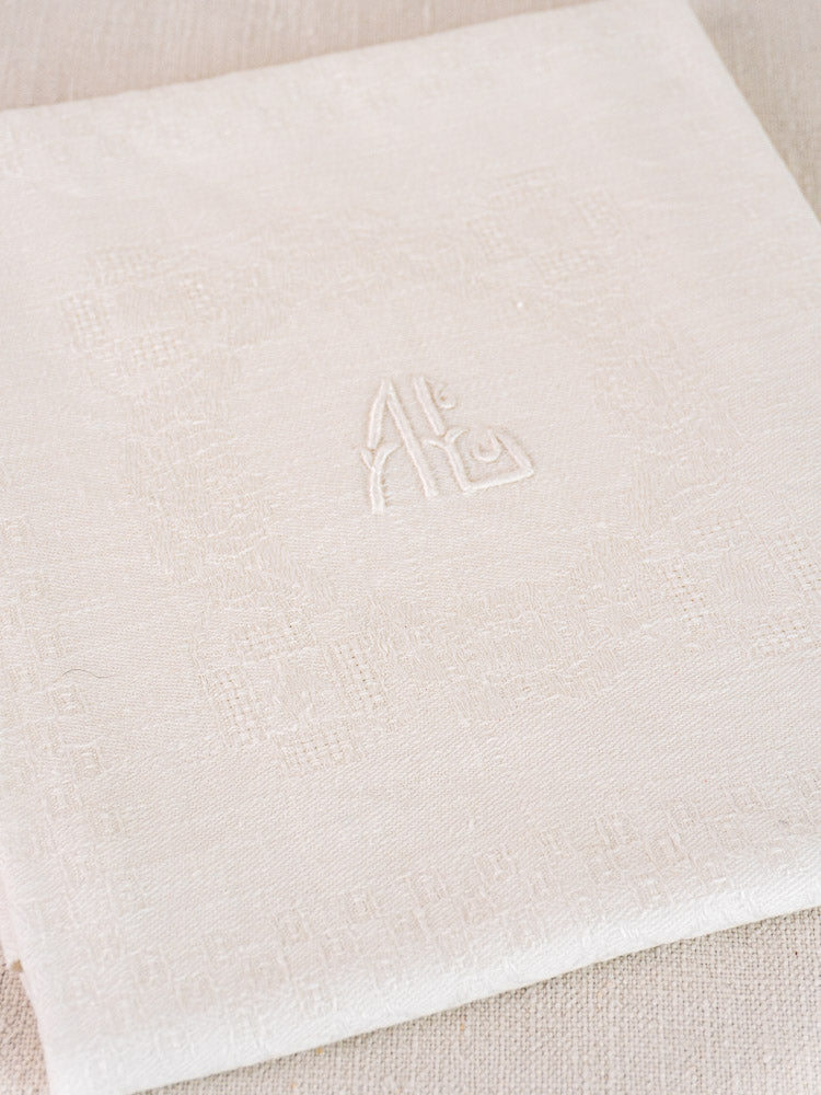 Folded antique French damask linen napkin with elegant monogram, light color on textured surface.