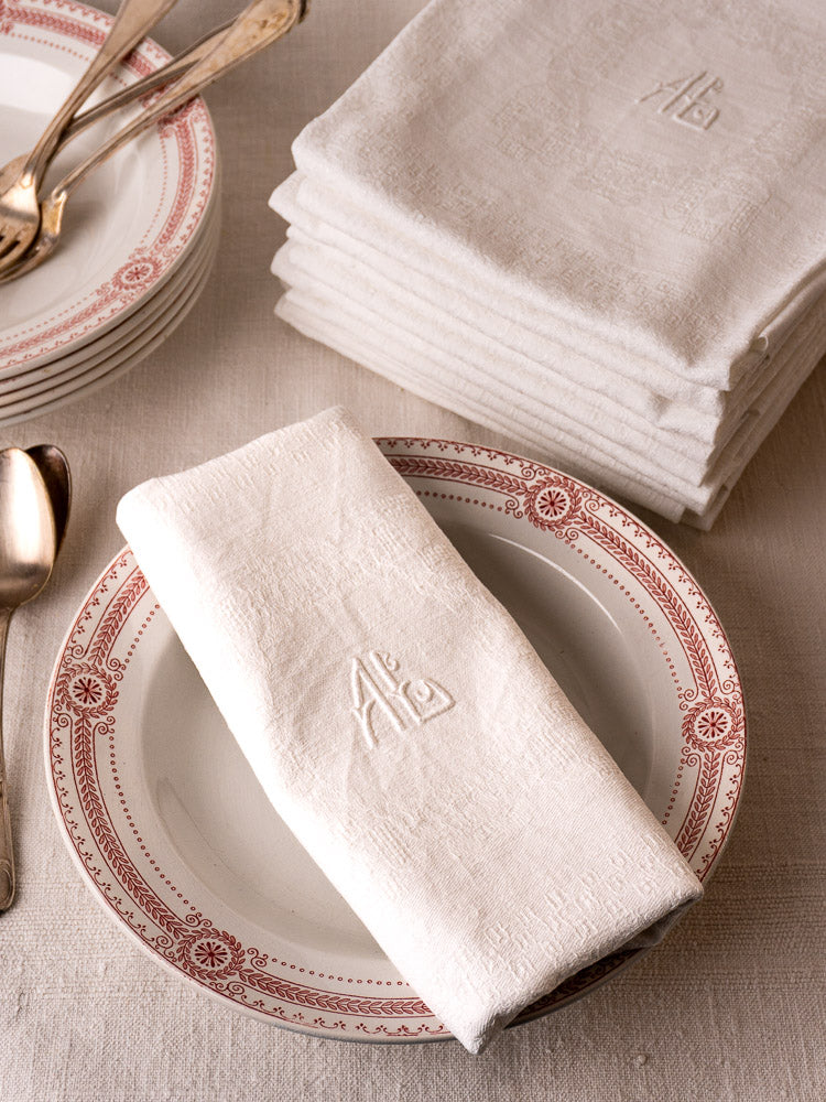 Set of 12 monogrammed antique French damask linen napkins beside plate, vintage silverware, red-patterned dishes on beige tablecloth.