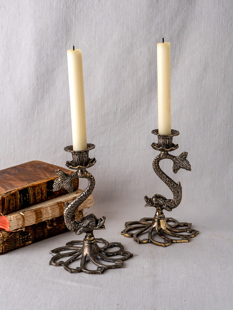 Pair of French antique silvered metal dolphin candlesticks with ornate bases on white fabric background.