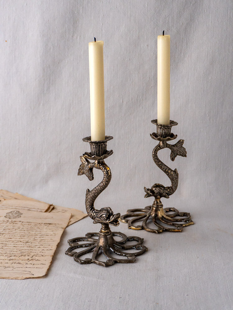 Pair of French antique silver dolphin candlesticks with tall cream candles on neutral fabric, vintage papers left.