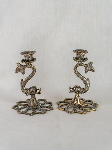 Pair of French antique silvered metal dolphin candlesticks with ornate bases on white fabric background.