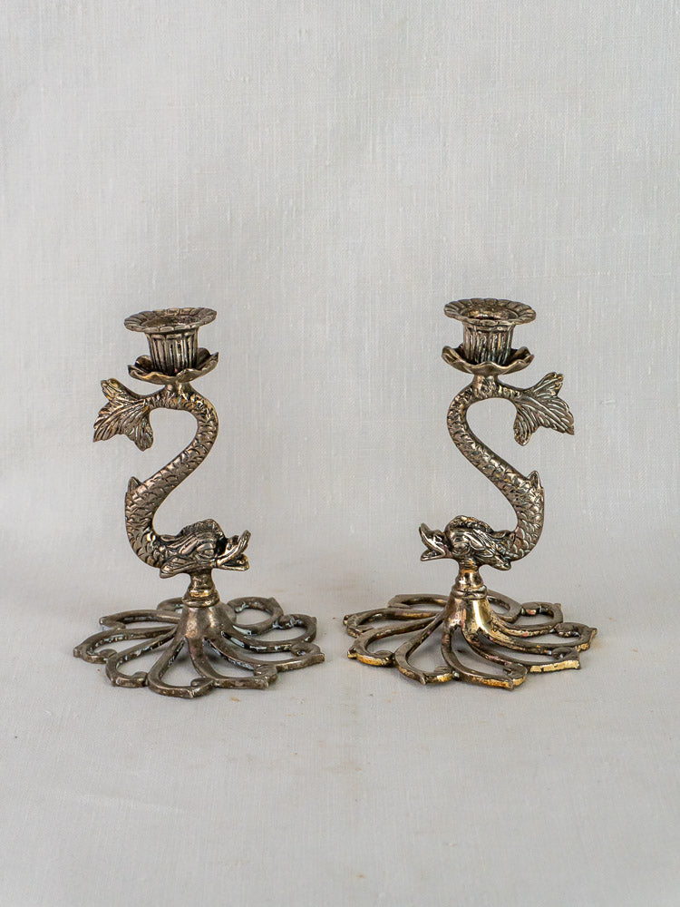 Pair of French antique silvered metal dolphin candlesticks with ornate bases on white fabric background.