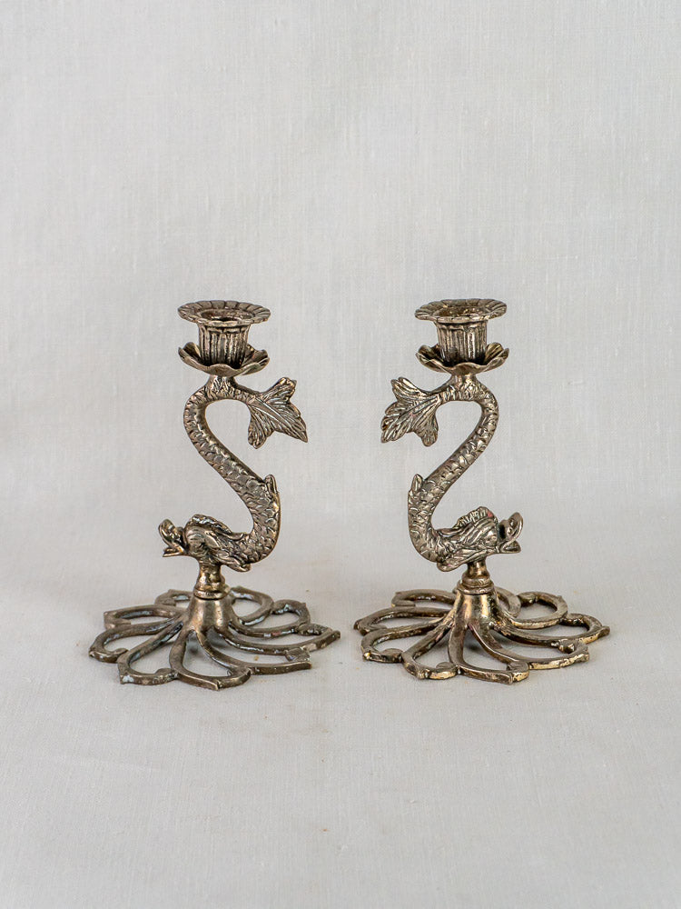 Pair of antique French dolphin candlesticks in silvered metal with intricate detailing on white fabric.