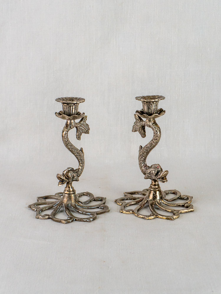 Ornate French silvered metal dolphin candlesticks with sculpted bases on a light fabric background.