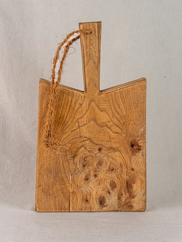 Mid-19th century French elm cutting board with rope handle and visible grain, displayed upright against light fabric.