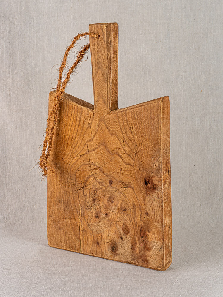 Antique French elm cutting board with rope handle, rectangular form, visible grain; set against neutral fabric backdrop.