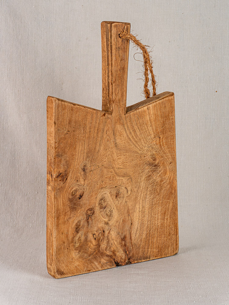 Antique French elm cutting board with rope handle, mid 19th century, vintage wood grain, light gray fabric backdrop.