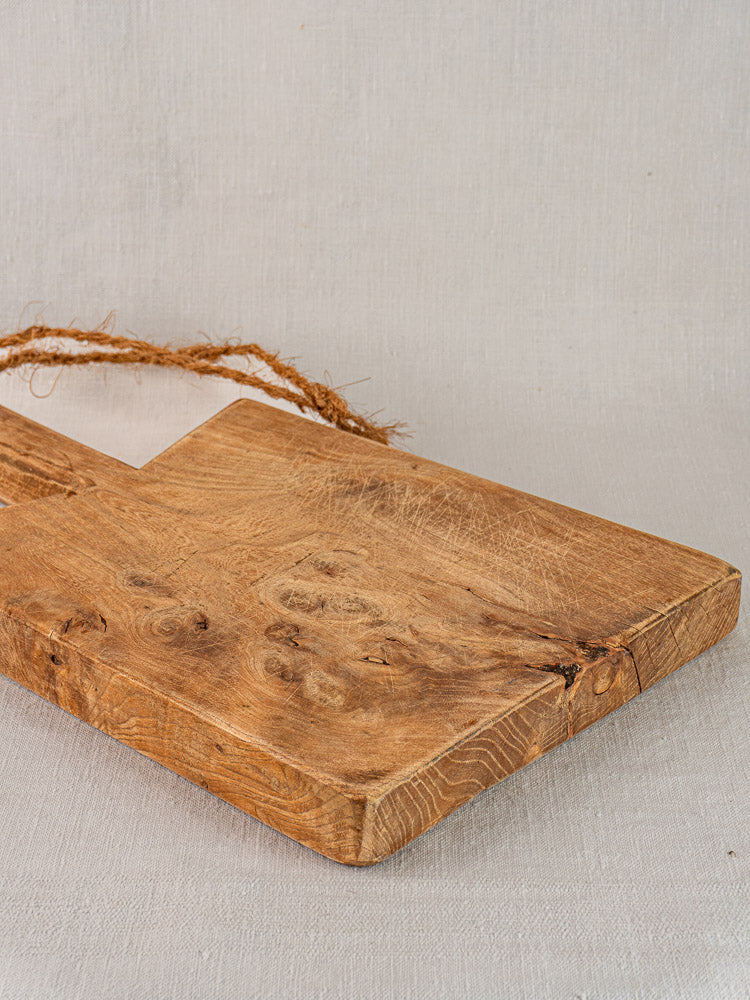 Antique French elm cutting board with rope handle, mid 19th century, displayed on a light fabric surface.