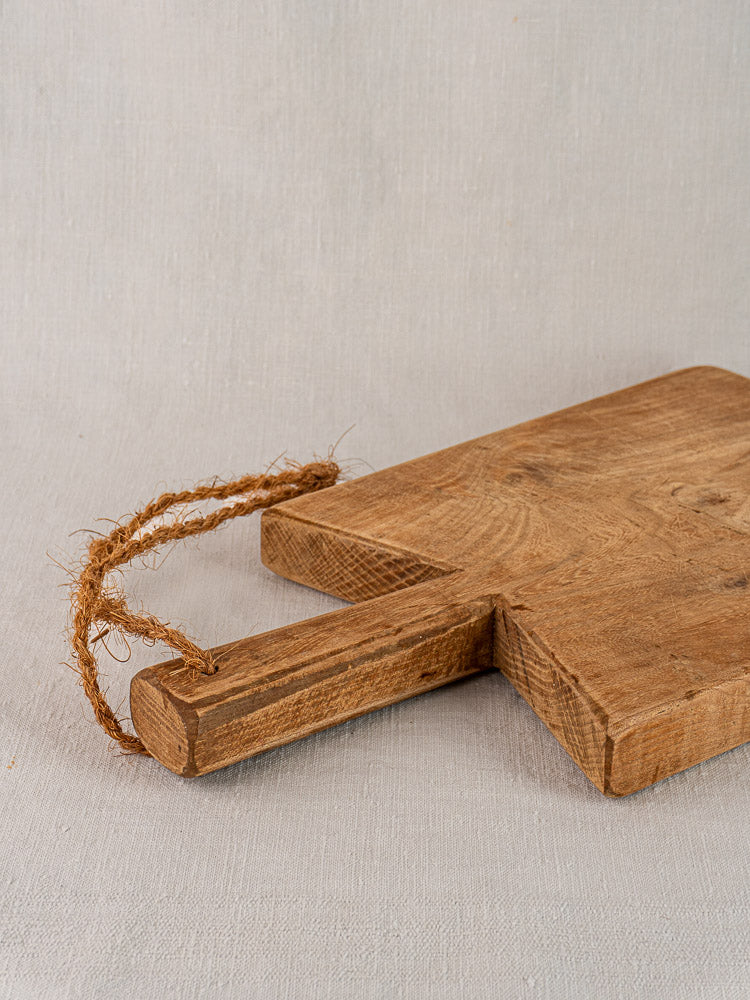 Antique French elm cutting board with rope handle, mid-19th century, on light gray fabric surface.