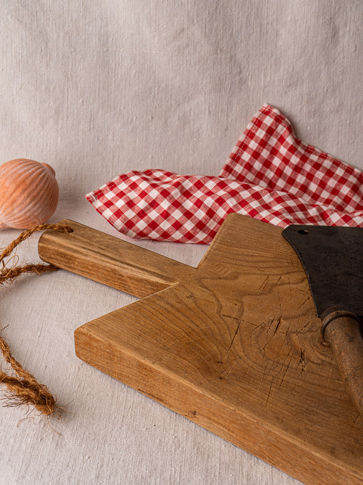 Mid-19th century French elm cutting board with rope handle on light fabric, shown with cleaver, checkered cloth, and seashell.