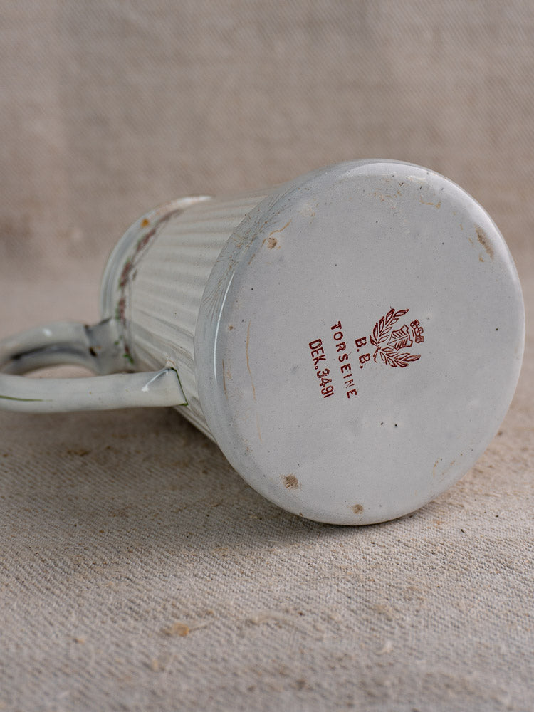 White ceramic mug on textured fabric, underside showing red text with laurel wreath motif, French vintage style.