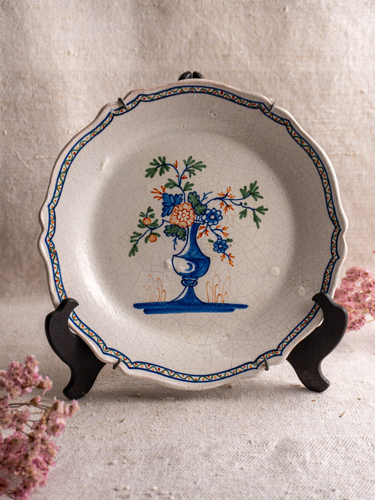 Antique French faience plate with scalloped edge, hand-painted floral vase, on black stand with pink blooms and beige fabric backdrop.