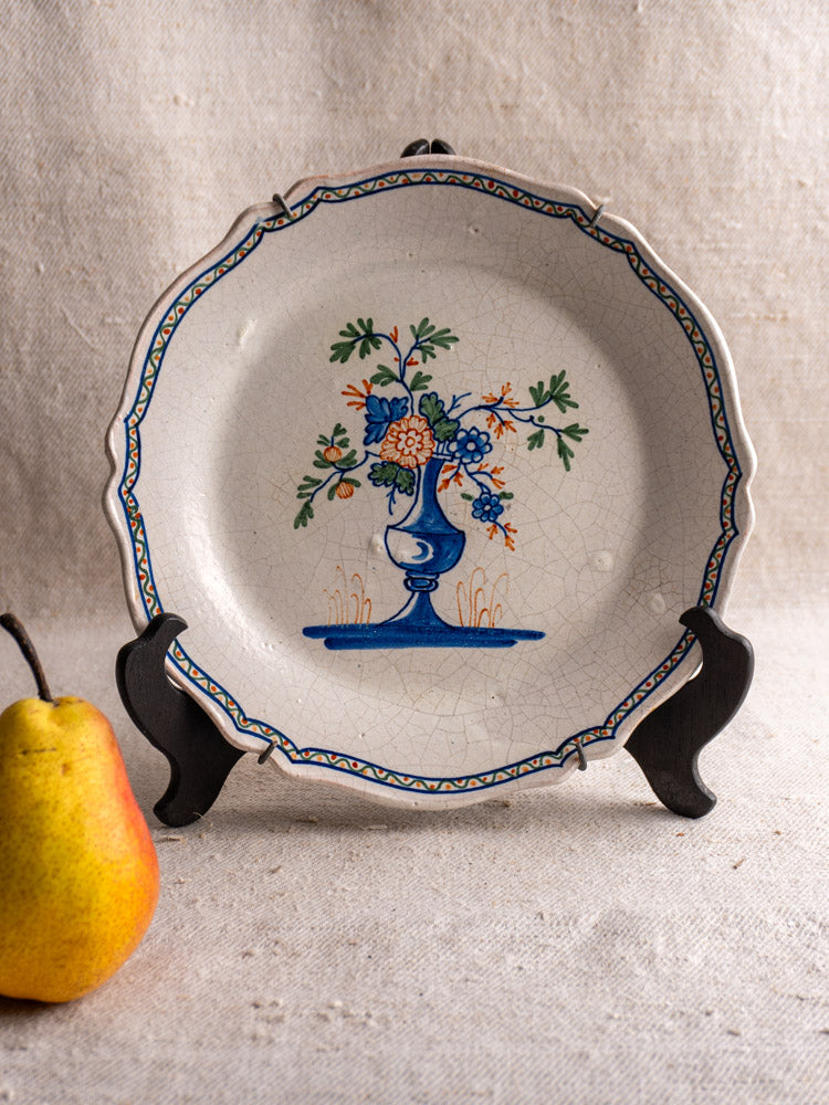 Antique French faience plate with hand-painted vase bouquet, blue and orange accents, on black stand; pear on beige textured fabric.